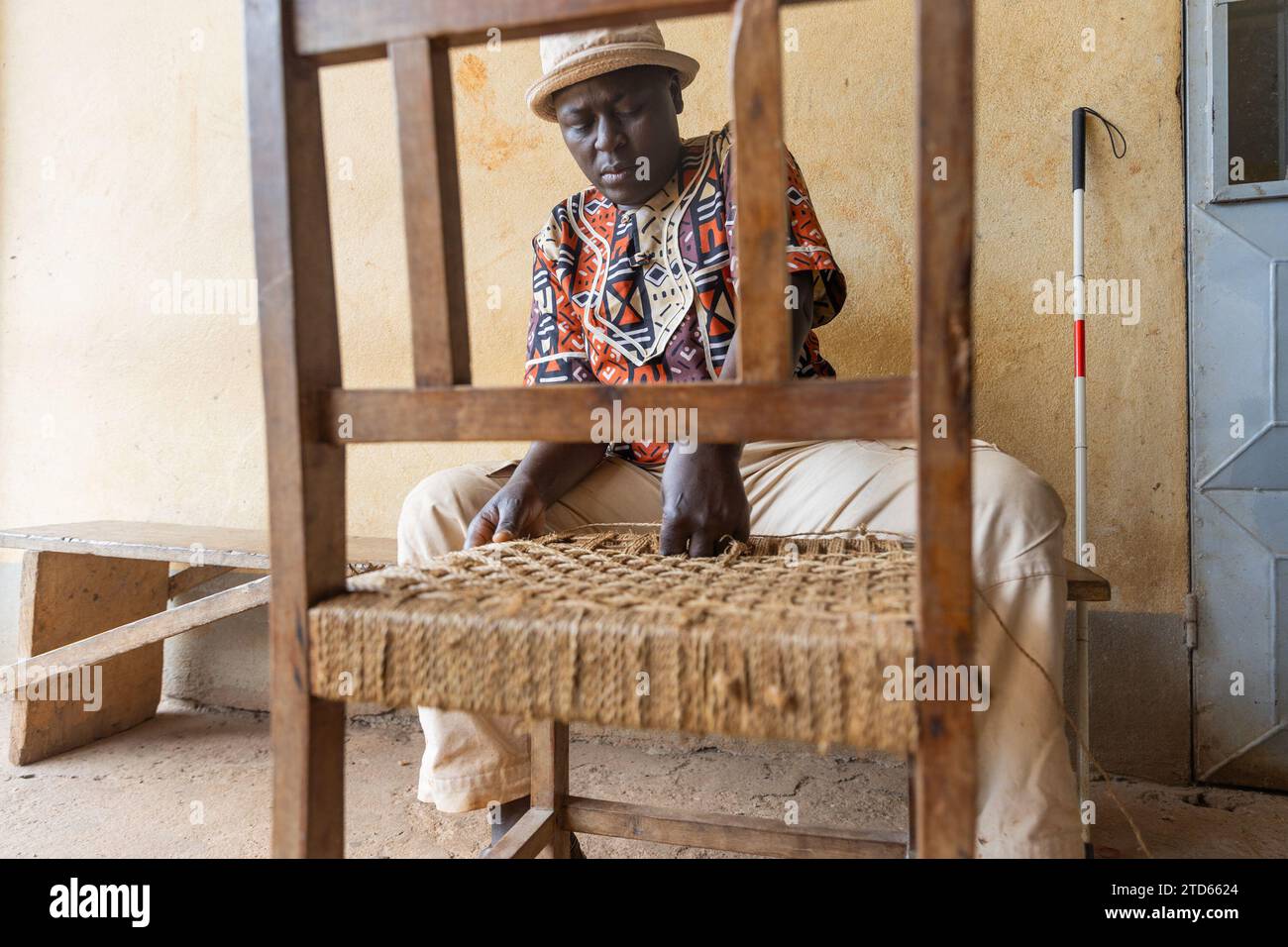 Lavoro di primo piano di tessitura di una sedia di legno da parte di un uomo africano cieco con cappello sulla testa. Foto Stock