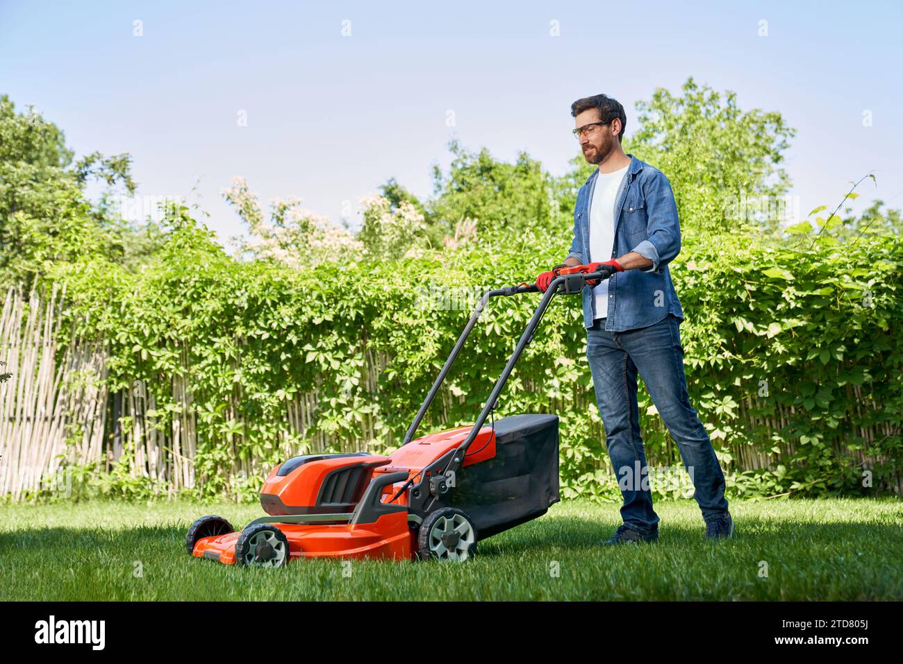 Bellissimo giardiniere maschile in guanti che rifinisce erba con un rasaerba elettrico in giardino di giorno. Vista laterale di un sorridente ragazzo barbuto con outfit in denim utilizzando il trimm per prato Foto Stock