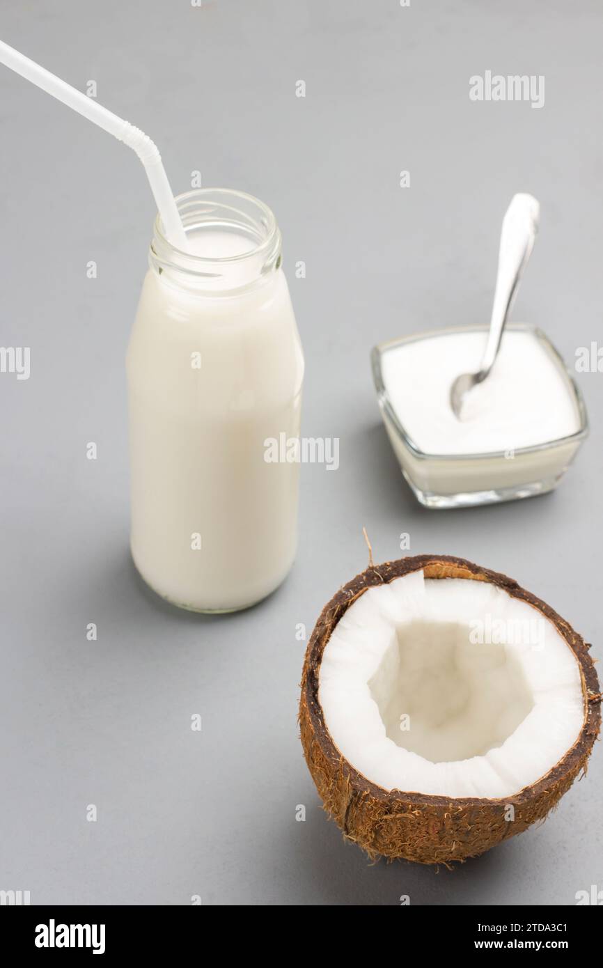 Mezzo cocco fresco. Bottiglia di latte di cocco, cannucce. Ciotola di yogurt, cucchiaio. Sfondo grigio Foto Stock