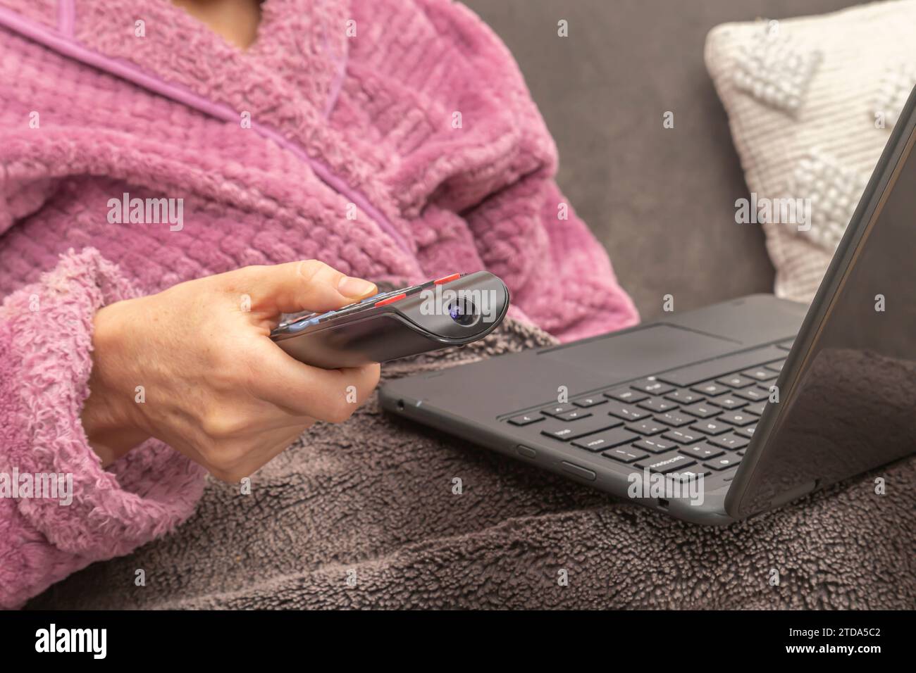 Donna seduta sul divano a casa con telecomando TV e laptop in grembo Foto Stock