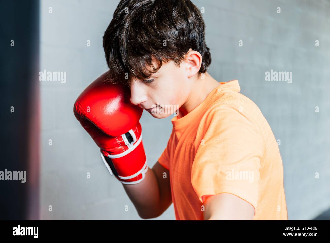 Foto orizzontale adolescente caucasico con t-shirt arancione e guanti da boxe rossi, che colpiscono la borsa di punzonatura in palestra. Concetto di sport, tempo libero. Foto Stock
