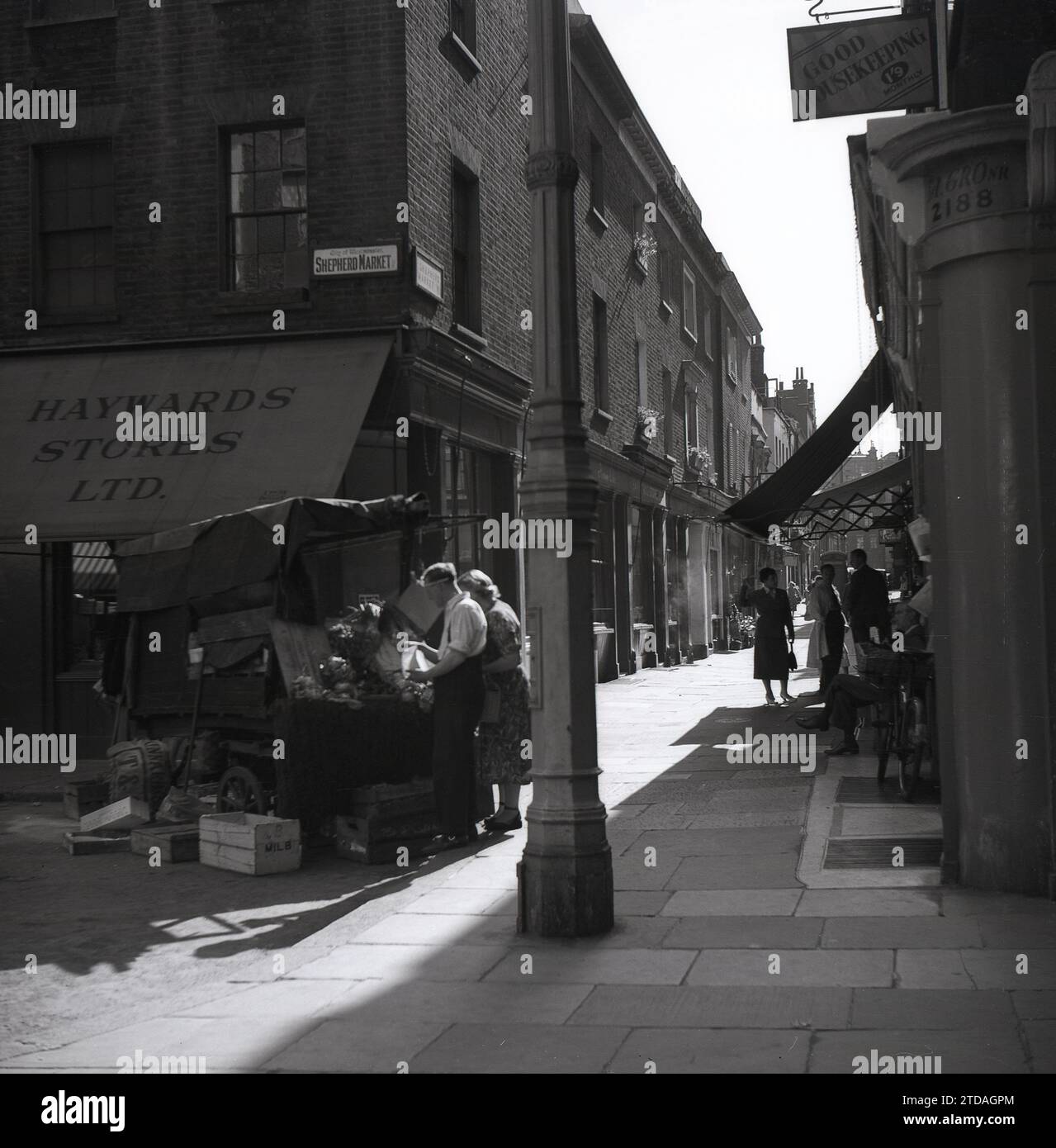 Anni '1950, storica, una donna che fa shopping con un commerciante di sesso maschile al Market Stall, Shepherd Market, a Mayfair, Londra, Inghilterra, REGNO UNITO. All'angolo del vicolo lastricato, Hayward Stores e un cartello per una buona casa magazzino a 1'9 al mese. Situato a Mayfair, in prossimità di Piccadilly, Shepherd Market è una parte affascinante del centro di Londra con la sua piccola piazza e le sue strette stradine laterali, essendo conosciuto come un "villaggio a Piccadilly". Foto Stock