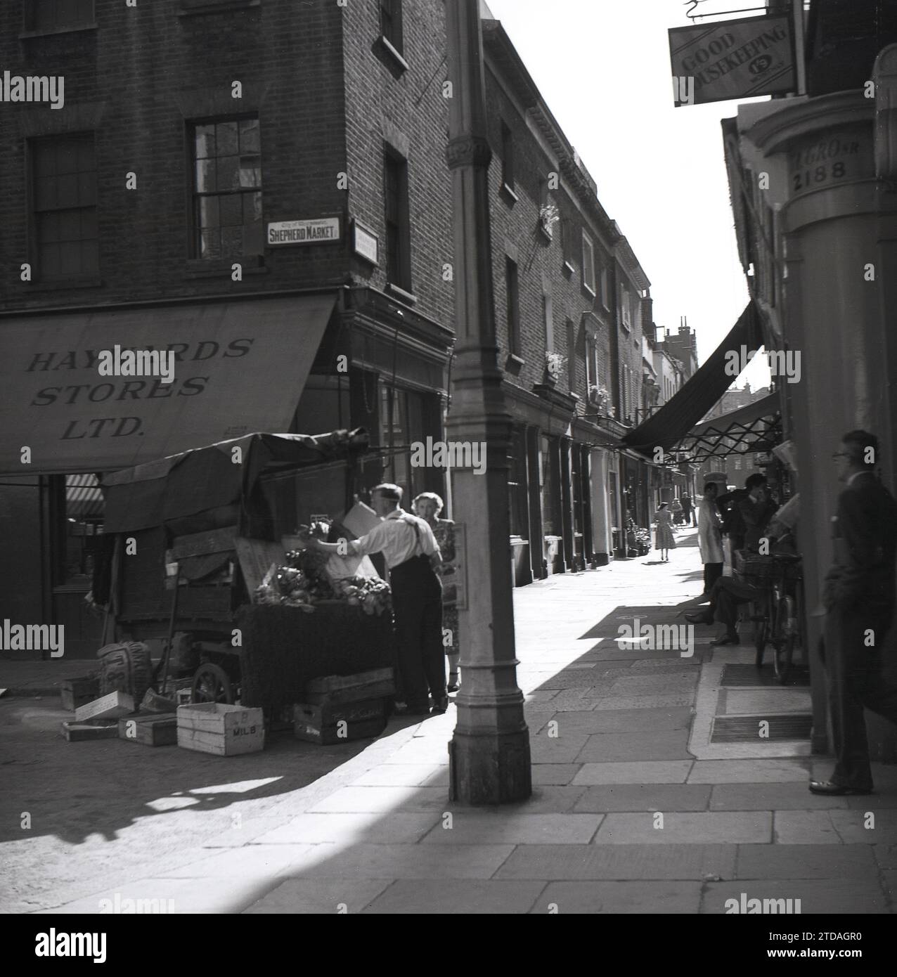 Anni '1950, storica, una donna che fa shopping con un commerciante di sesso maschile al Market Stall, Shepherd Market, a Mayfair, Londra, Inghilterra, REGNO UNITO. All'angolo del vicolo lastricato, Hayward Stores e un cartello per una buona casa magazzino a 1'9 al mese. Situato a Mayfair, in prossimità di Piccadilly, Shepherd Market è una parte affascinante del centro di Londra con la sua piccola piazza e le sue strette stradine laterali, essendo conosciuto come un "villaggio a Piccadilly". Foto Stock