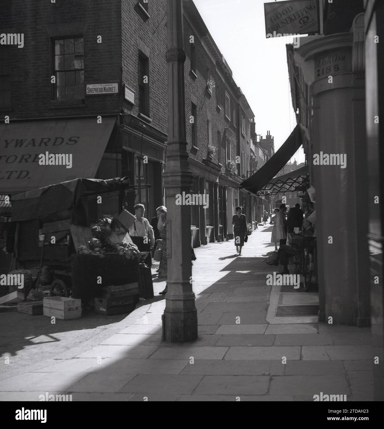 Anni '1950, storica, una donna che fa shopping con un commerciante di sesso maschile al Market Stall, Shepherd Market, a Mayfair, Londra, Inghilterra, REGNO UNITO. All'angolo del vicolo lastricato, Hayward Stores e un cartello per una buona casa magazzino a 1'9 al mese. Situato a Mayfair, in prossimità di Piccadilly, Shepherd Market è una parte affascinante del centro di Londra con la sua piccola piazza e le sue strette stradine laterali, essendo conosciuto come un "villaggio a Piccadilly". Foto Stock