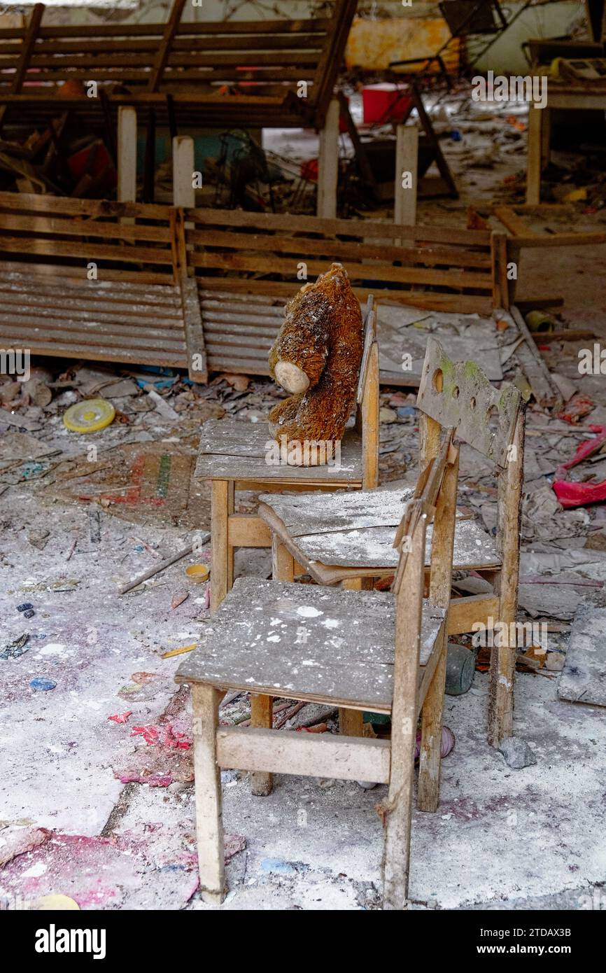 Un orsacchiotto si siede su una sedia di legno tra detriti e mobili abbandonati. Peluche sporco sulla sedia. Un orsacchiotto senza testa in un asilo abbandonato Foto Stock