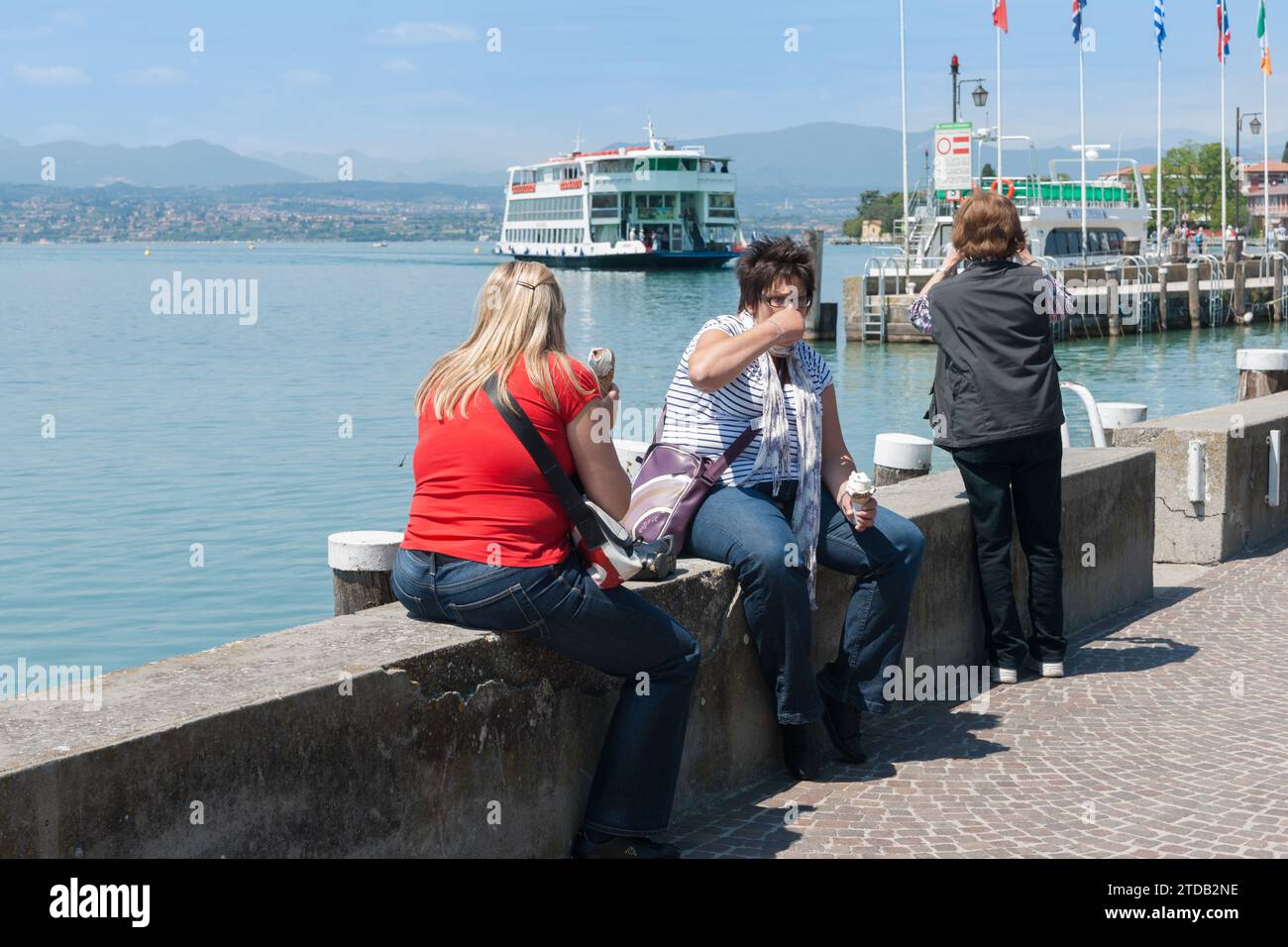 Sermione Italia - 9 maggio 2011; due donne con gelato sedute su una parete bassa sul bordo del lago e una in piedi che scatta foto al molo e si avvicina al traghetto. Foto Stock