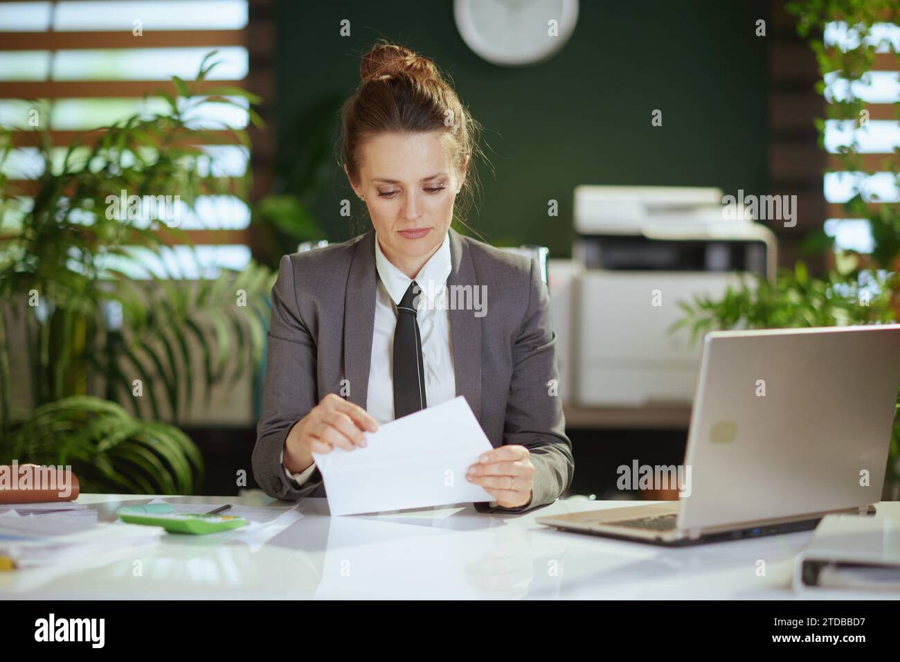 Nuovo lavoro. penosa moderna 40 anni donna impiegata in un moderno ufficio verde in tuta da lavoro grigia con lettera di apertura del laptop. Foto Stock