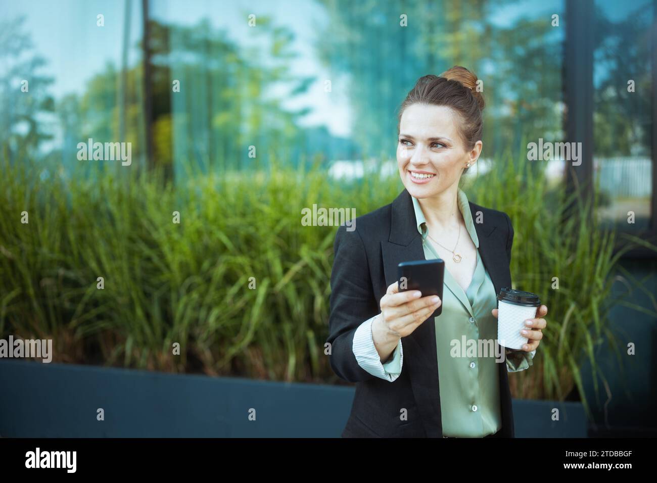 lavoratrice moderna sorridente vicino al business center in una giacca nera con una tazza di caffè utilizzando lo smartphone. Foto Stock