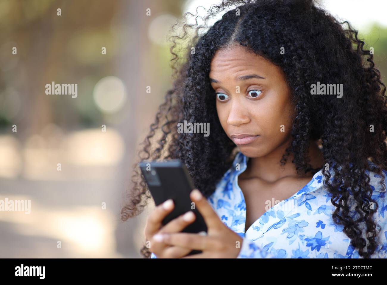 Donna nera perplessa che guarda i nuovi smartphone online in un parco Foto Stock