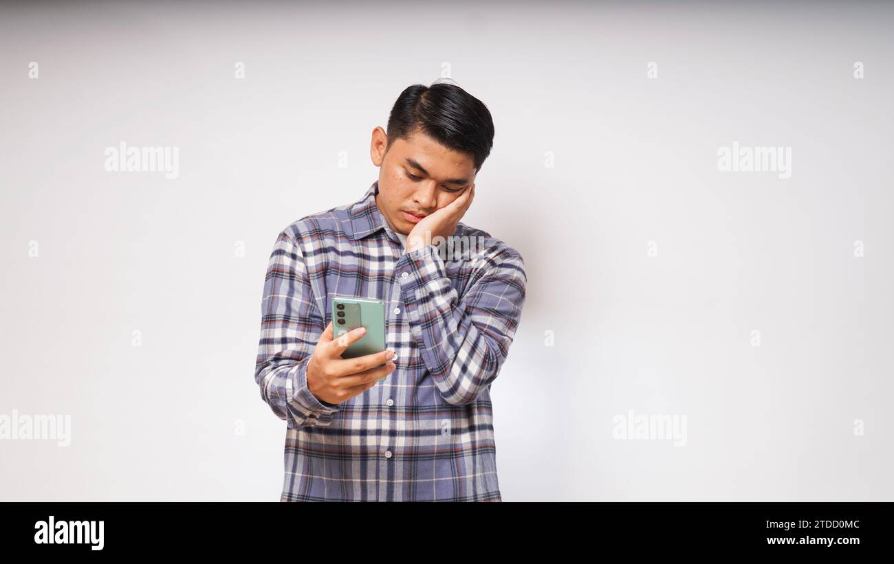 Giovane asiatico che tiene il suo telefono cellulare con un'espressione triste e confusa su sfondo bianco. foto studio Foto Stock