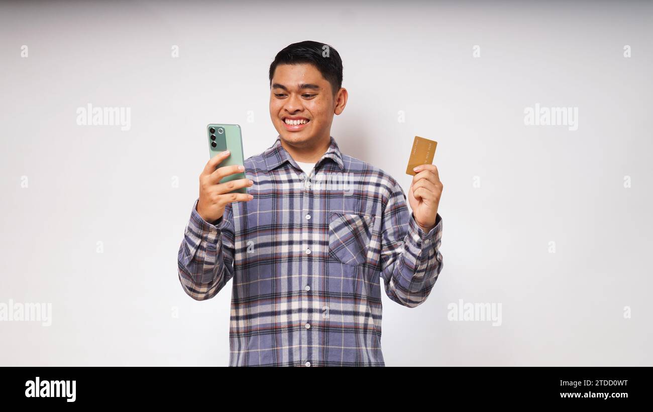 Ritratto di un giovane asiatico soddisfatto con smartphone e carta di credito, inserimento dati, shopping online, maglietta casual. Monolocali interni Foto Stock