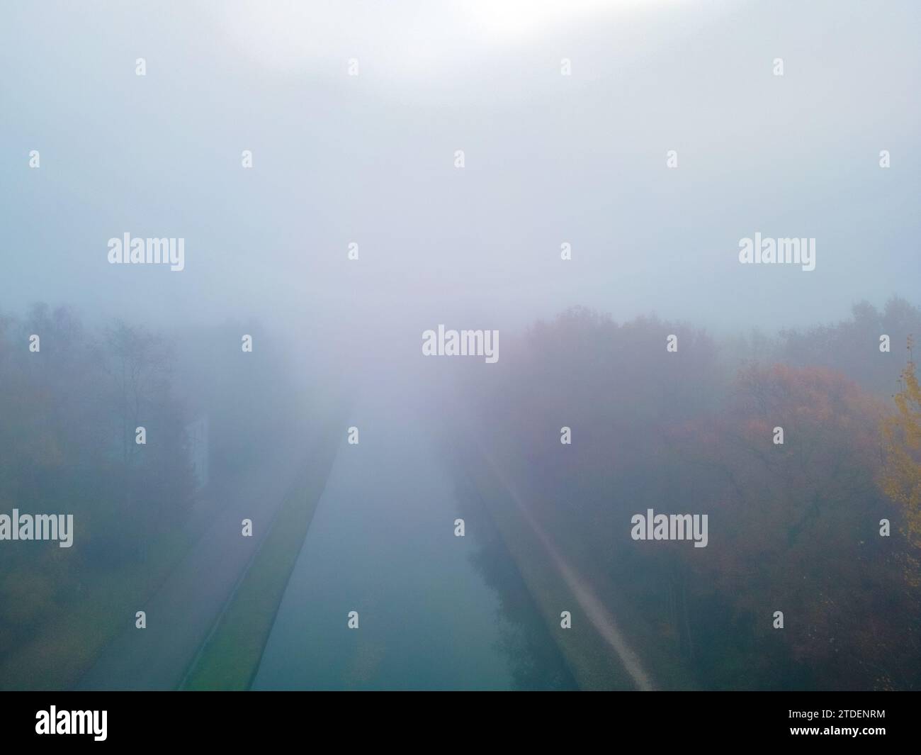 In questa fotografia, un canale scorre dritto attraverso la cornice, fiancheggiato da alberi su entrambi i lati nel pieno flusso dell'autunno. I colori del fogliame sono attenuati da una fitta e avvolgente nebbia che ammorbidisce l'intera scena, creando una sensazione di mistero e quiete. La nebbia oscura qualsiasi caratteristica distinta nella distanza, concentrando l'attenzione dello spettatore sulla simmetria vicina del canale e degli alberi. L'immagine cattura l'essenza di una tranquilla mattinata autunnale in cui il mondo è coperto e l'aria è spessa di nebbia. Canale fiancheggiato da alberi autunnali nella fitta nebbia. Foto di alta qualità Foto Stock