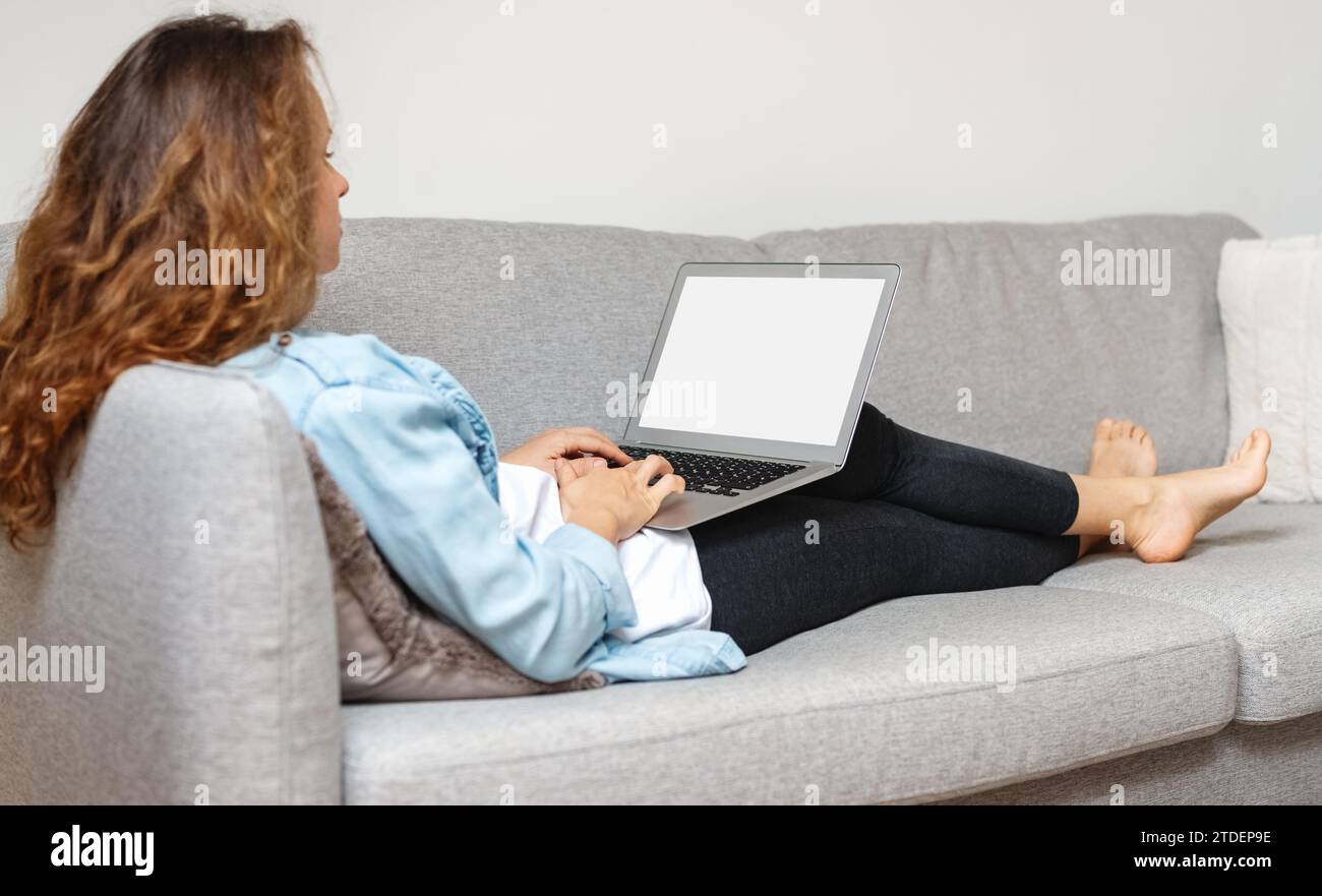 Vista posteriore della donna seduta sul divano e al lavoro sul computer portatile. Schermo vuoto del laptop. Foto Stock