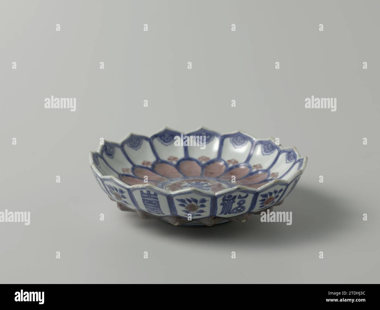 Piatto, anonimo, c. 1672 Scala di porcellana modellata a forma di loto, dipinto in blu e rosso. Sul piatto un medaglione con il carattere "dovrebbe" circondato da una banda con motivo Ruyi; intorno a un bordo petali modellati che si sovrappongono a un secondo bordo petali appuntiti con motivo Ruyi; le foglie del bordo superiore hanno rami floreali alternati all'esterno o il carattere "dovrebbe" o "fu", entrambi volti felici. Sul fondo una sala di otto caratteri segna 'Kangxi Renzi Zhonghetang Zhi'. Frammenti multipli nel bordo; i punti della fila inferiore di petali all'esterno sono danneggiati. Sottosmalto blu W Foto Stock