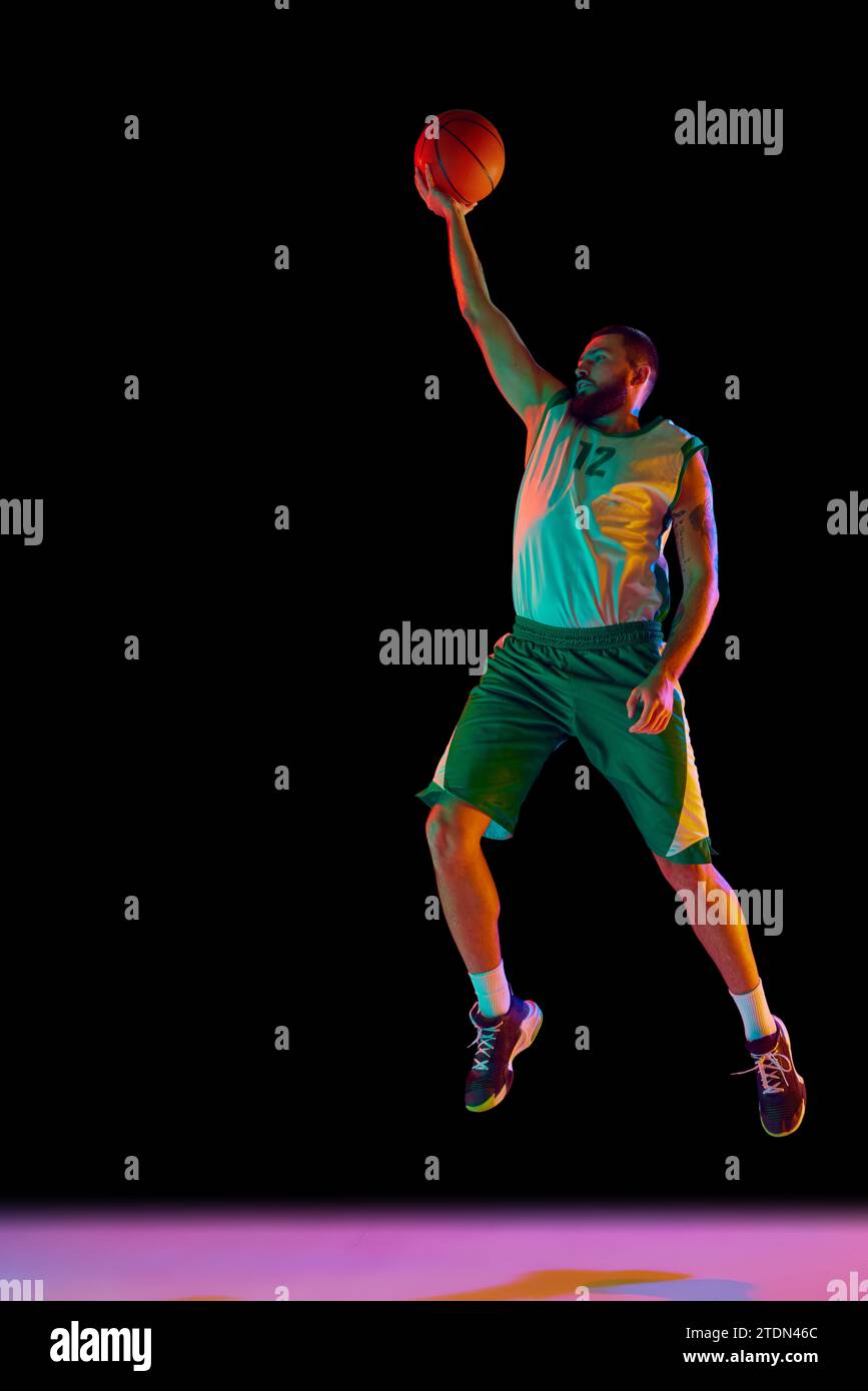 Ritratto dinamico di uomo atletico, giocatore professionista di basket che perfeziona le proprie abilità con dribbling di precisione su sfondo nero in luce neon. Foto Stock