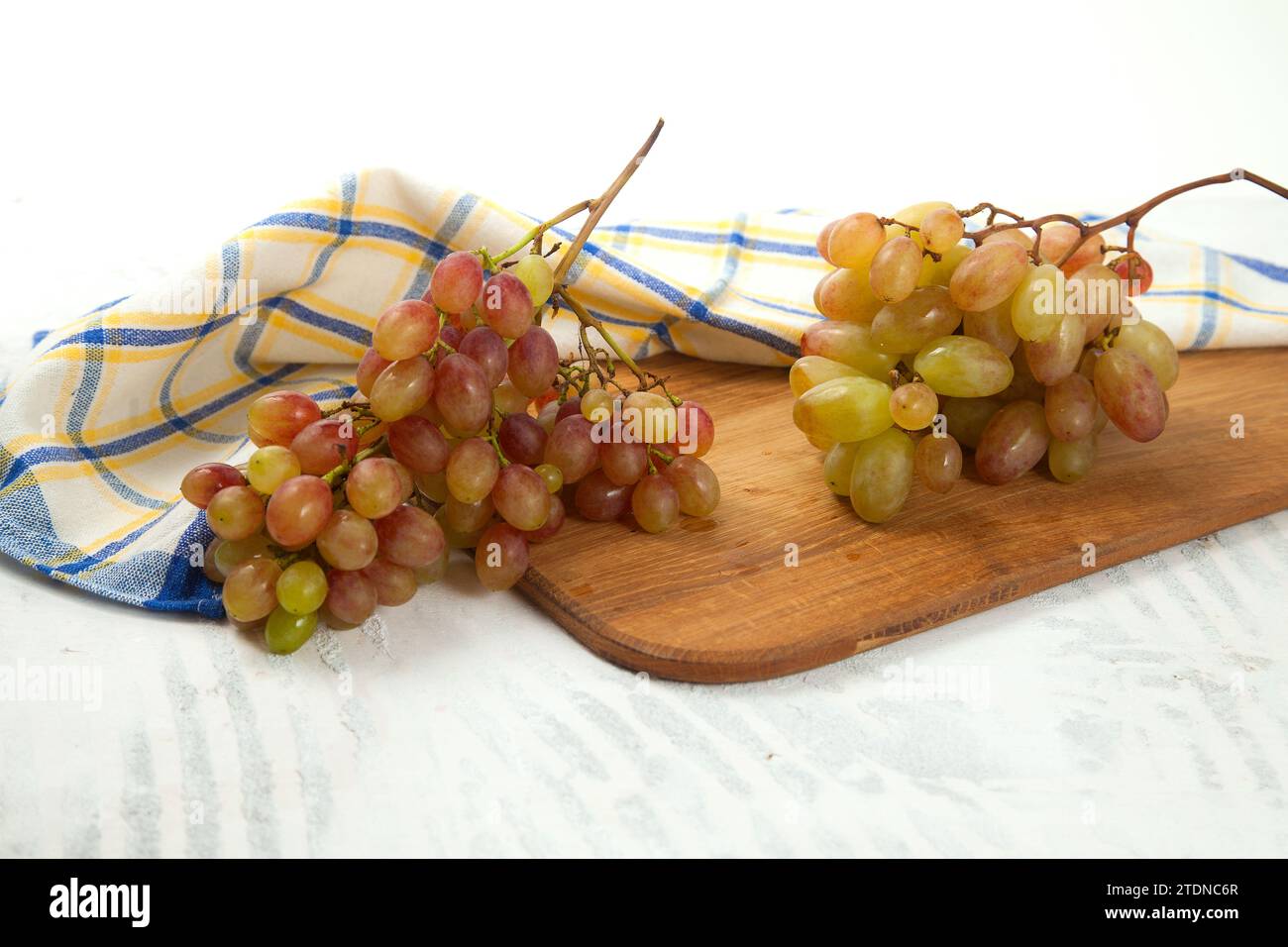 Tagliere con grande mazzo di uva rosa matura su bianco. Uva da vino rosa con asciugamano da cucina blu su fondo di legno bianco. Foto Stock