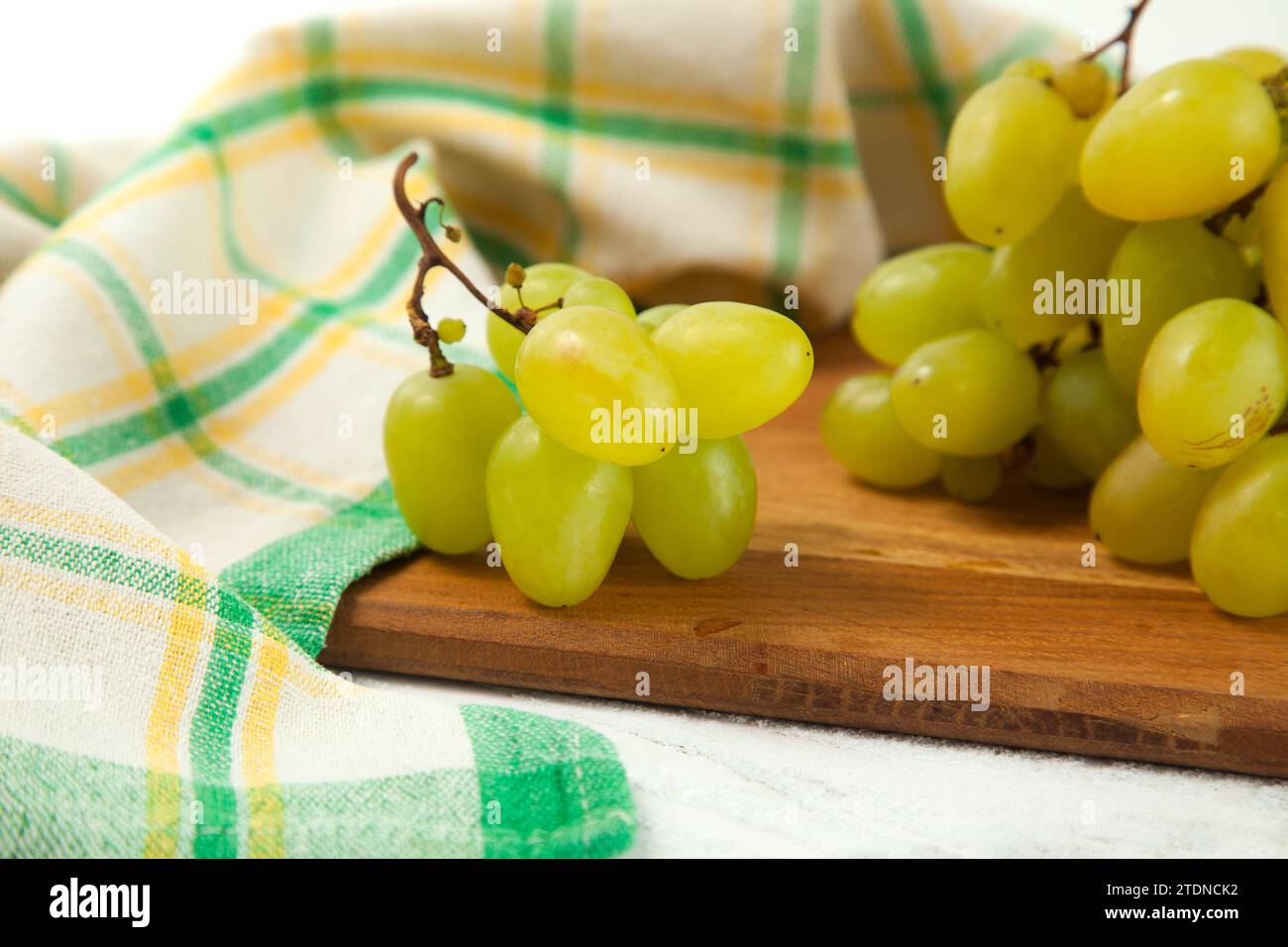 Tagliere con grande mazzo di uva verde matura su bianco. Uva da vino bianco con asciugamano da cucina verde su fondo di legno bianco. Foto Stock