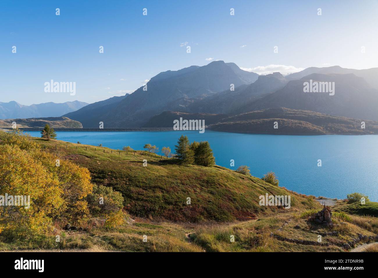 Monte Cenis (Moncenisio) lago e montagne, cielo blu e luce soffusa, sponda con alberi in autunno, diga e acqua limpida Foto Stock