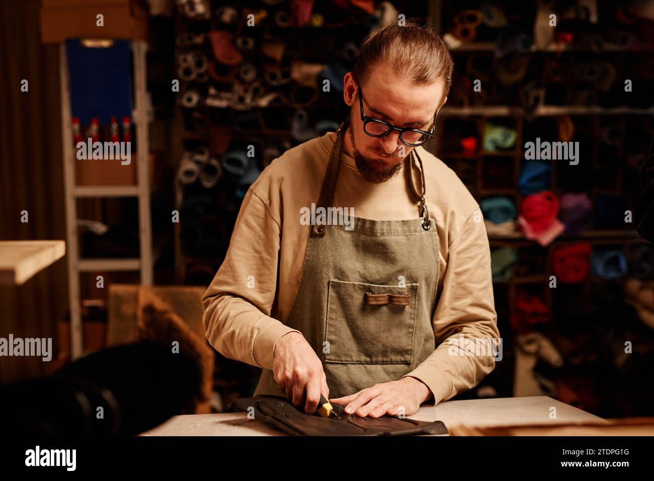 Giovane esperto di calzatura professionale in occhiali da vista e abbigliamento da lavoro che taglia i pezzi della suola sul posto di lavoro mentre si trova di fronte alla fotocamera Foto Stock
