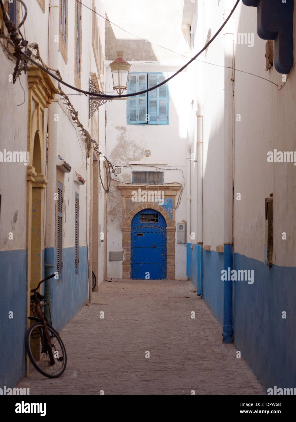 Strada stretta blu e bianca con bicicletta e porta e finestra blu in legno nella medina della città di Essaouira, Marocco. 19 dicembre 2023 Foto Stock