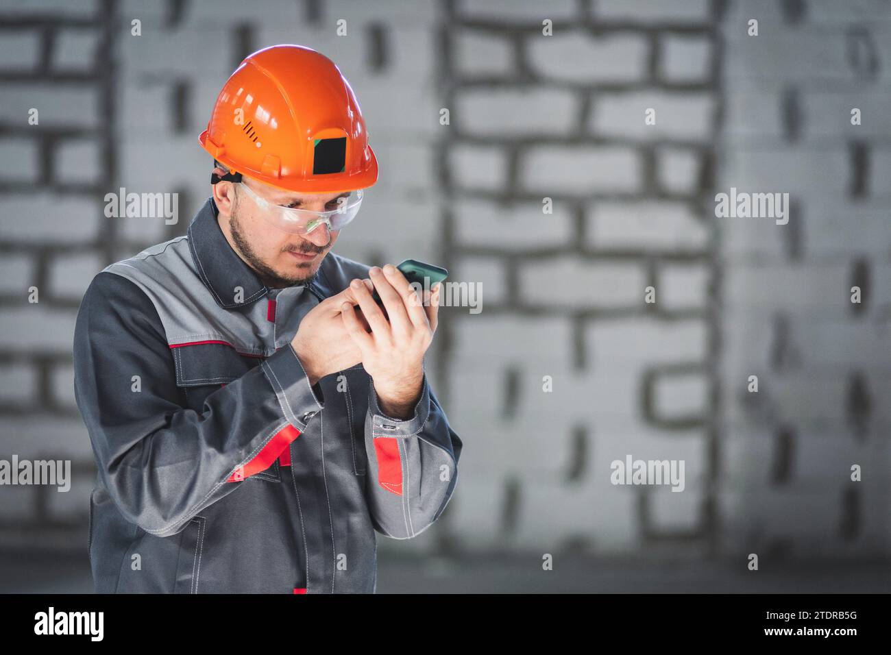 Il costruttore maschio caucasico in tute e casco utilizza lo smartphone come radio per la comunicazione in cantiere Foto Stock