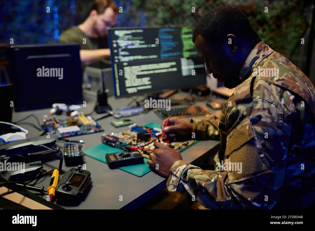 Giovane serio ufficiale di sicurezza afroamericano in uniforme mimetica che ripara il circuito stampato mentre è seduto sul posto di lavoro Foto Stock