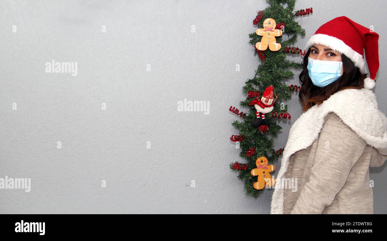 Donna latina con maschera protettiva, maglione e cappello di natale su sfondo grigio con decorazione natalizia, nuovo normale Foto Stock