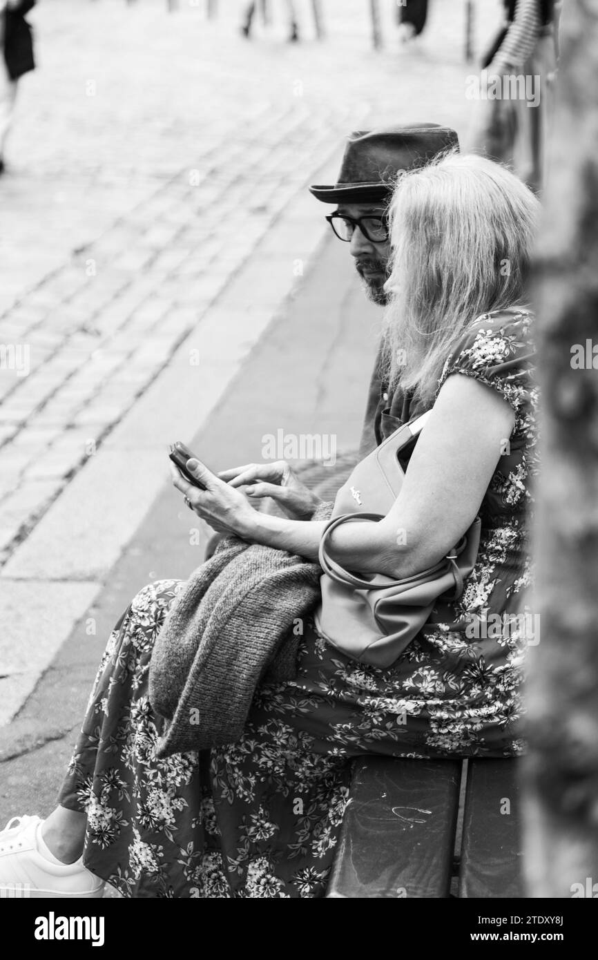 Coppia seduta su una panchina in strada a guardare il cellulare, Parigi, Francia Foto Stock