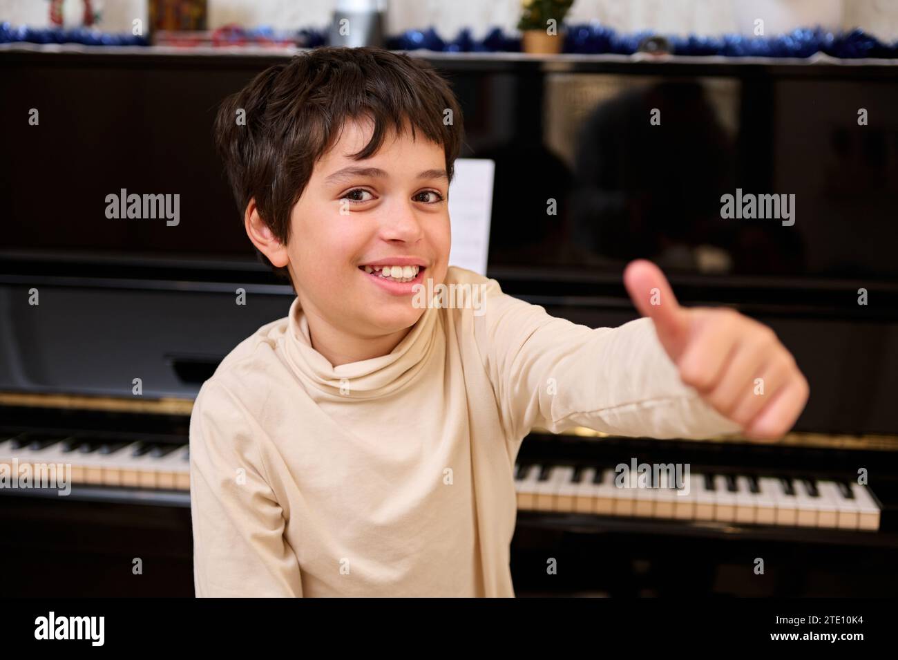 Allegro scolaro dell'età elementare che guarda la fotocamera e sorride mentre guarda il piano durante la lezione di musica. Ragazzo di talento carino e felice, un Foto Stock