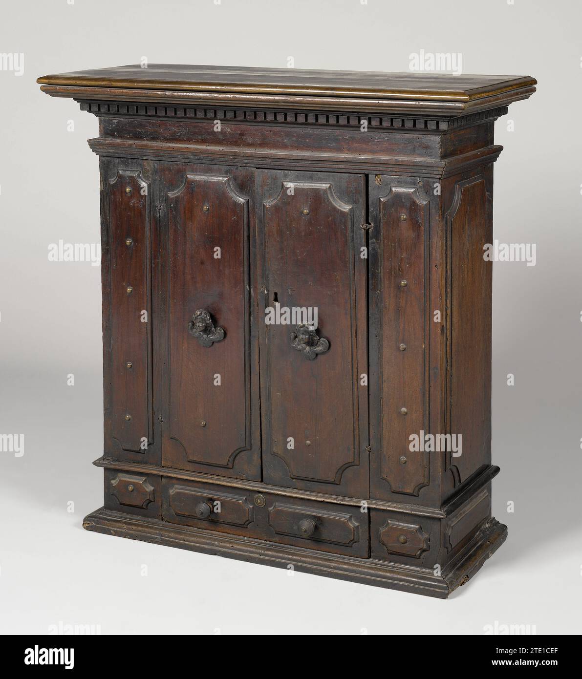 Armadio a parete nuthout, anonimo, 1500 - 1600 armadio a parete in noce, poggiato su un fondo profilato. L'armadietto è dotato di un cassetto nella parte inferiore, al di sopra del quale sono presenti due sportelli, tutti pannelli con angoli smussati. Sia il cassetto che gli sportelli sono fiancheggiati da un pannello simile più stretto. I pannelli da e accanto al cassetto sono dotati di un pulsante di disegno. Le porte mostrano pulsanti in bronzo sotto forma di teste in cherubino alato al centro; nella serratura una vecchia chiave con maniglia aperta. La parte superiore ha molti profili e sporge. Legno di Bologna (materiale vegetale). noce (legno duro). Armadio a parete in bronzo (metallo) in noce, Foto Stock