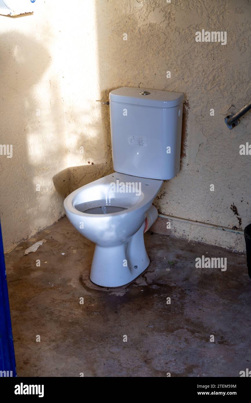 Un WC in ceramica con cisterna in ceramica in una camera sporca senza sedile. Potrai vedere i raggi del sole e un corrimano in camera. Il bagno sì Foto Stock