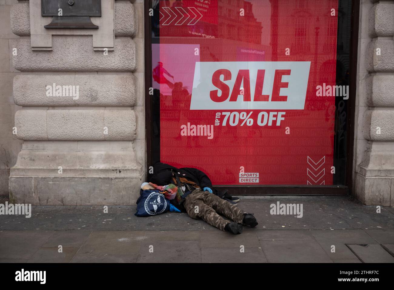 Un senzatetto si trova sulla strada di fronte a un cartello di sconto del 70% sul Piccadilly Circus di Londra, durante il periodo natalizio, Inghilterra, Regno Unito Foto Stock