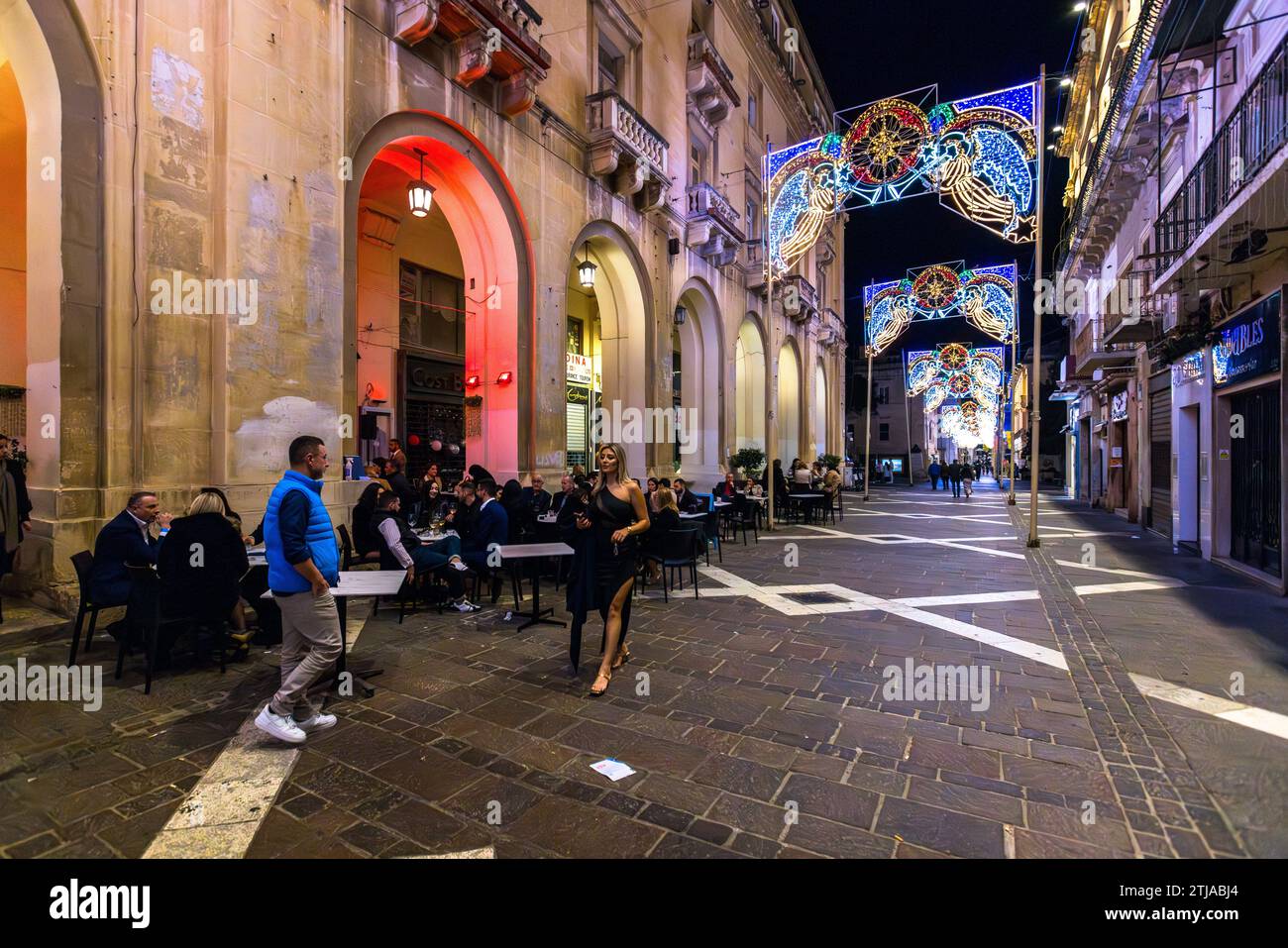 Le persone cenano e passeggiano sotto installazioni luminose natalizie a forma di stella in una strada ad arcate del centro storico di Valletta, Malta, dove le miti temperature di dicembre mantengono pieni i tavoli dei caffè all'aperto dopo il tramonto. Foto Stock