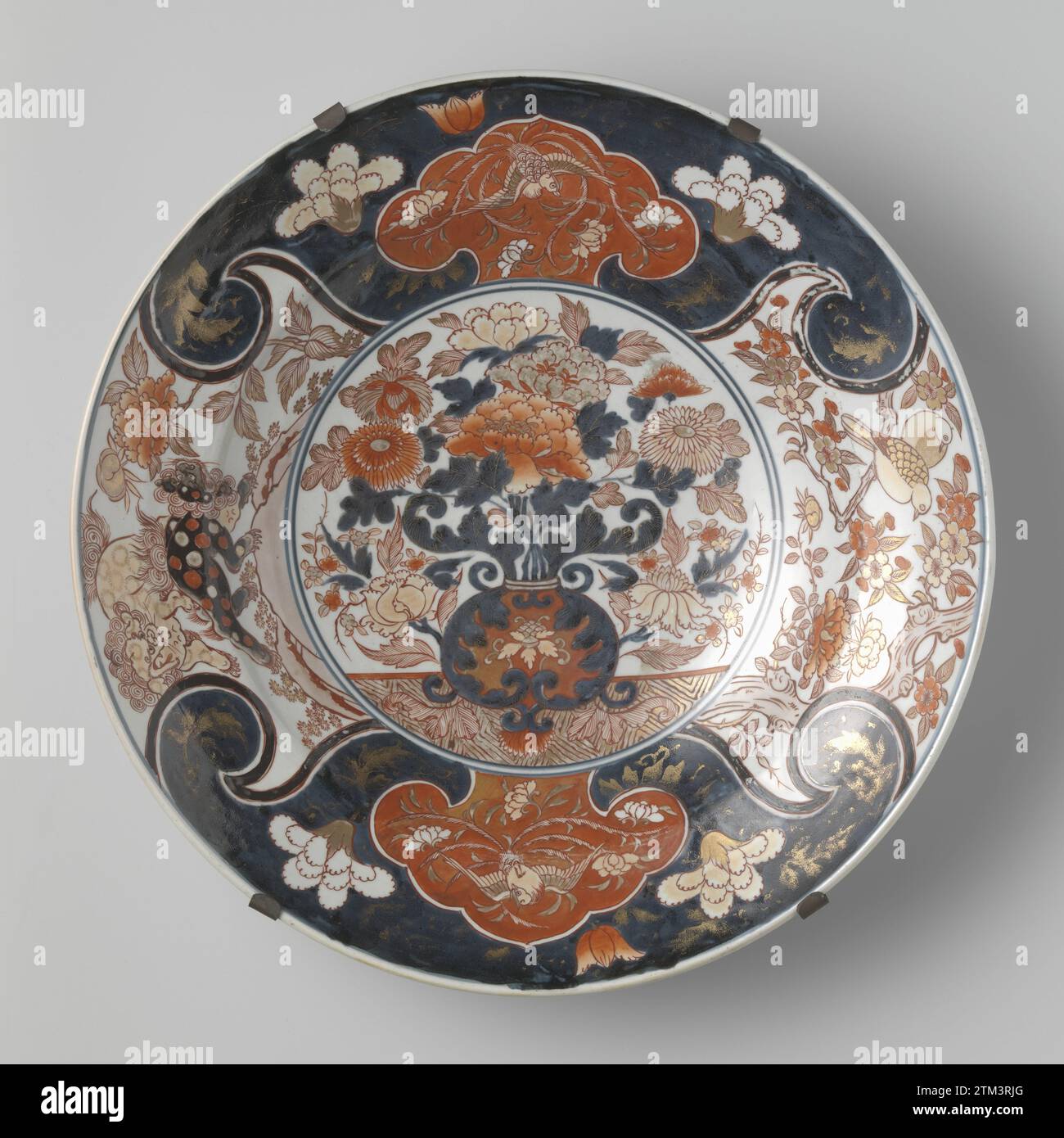 Piatto con vaso floreale, feng huangs, shishi and Birds, anonimo, c. 1700 - c. 1750 piattino in porcellana, dipinto in blu trasparente e rosso, nero e oro. Sul piatto un doppio medaglione con un vaso floreale con crisantemi e peonie. Il muro e il bordo sono divisi in quattro compartimenti con due shishi (cane da leone) in un paesaggio, due uccelli su un ramo di un albero di prunus e due volte un cartiglio a forma di Ruyi con un Fenghuang su uno sfondo blu con viti floreali. La piastra è intrappolata in una struttura metallica. Probabilmente un europeo (francese?) Imitazione dell'Imari giapponese. Europa Porcelai Foto Stock