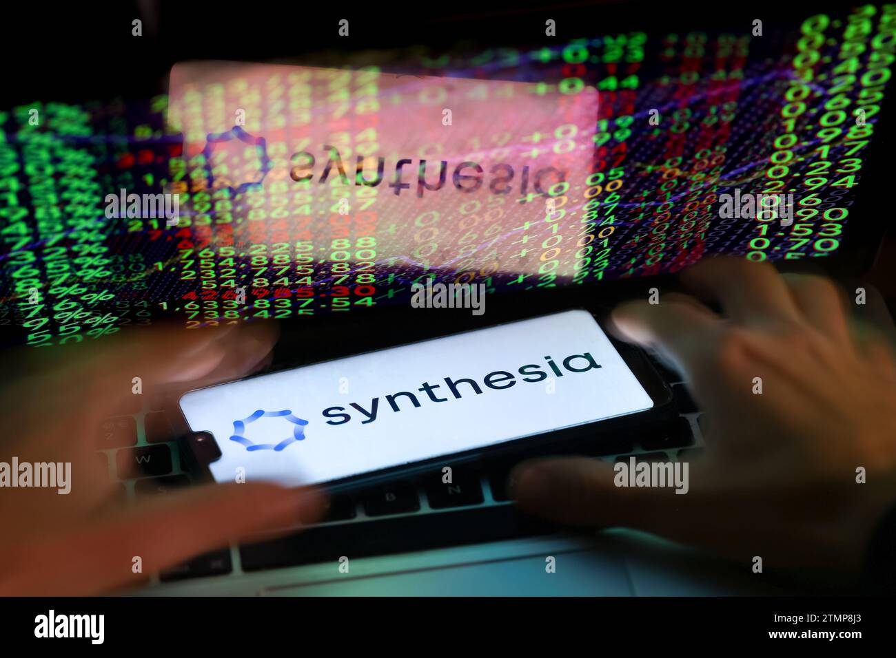 Cracovia, Polonia, 20 dicembre 2023. Questa fotografia mostra un logo dell'app Synthesia ai visualizzato su uno smartphone e mani umane che lavorano su un computer con i numeri di mercato azionario sullo sfondo. Foto Stock