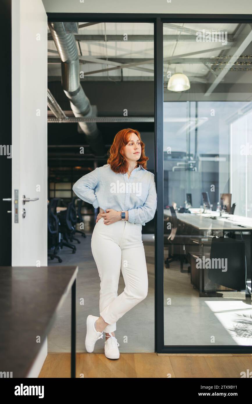 Una donna d'affari sicura di sé, redhead, si stupisce nel suo ufficio, raffigurando un dipendente dedicato nel settore tecnologico. Il suo aspetto trasuda fiducia e prof Foto Stock