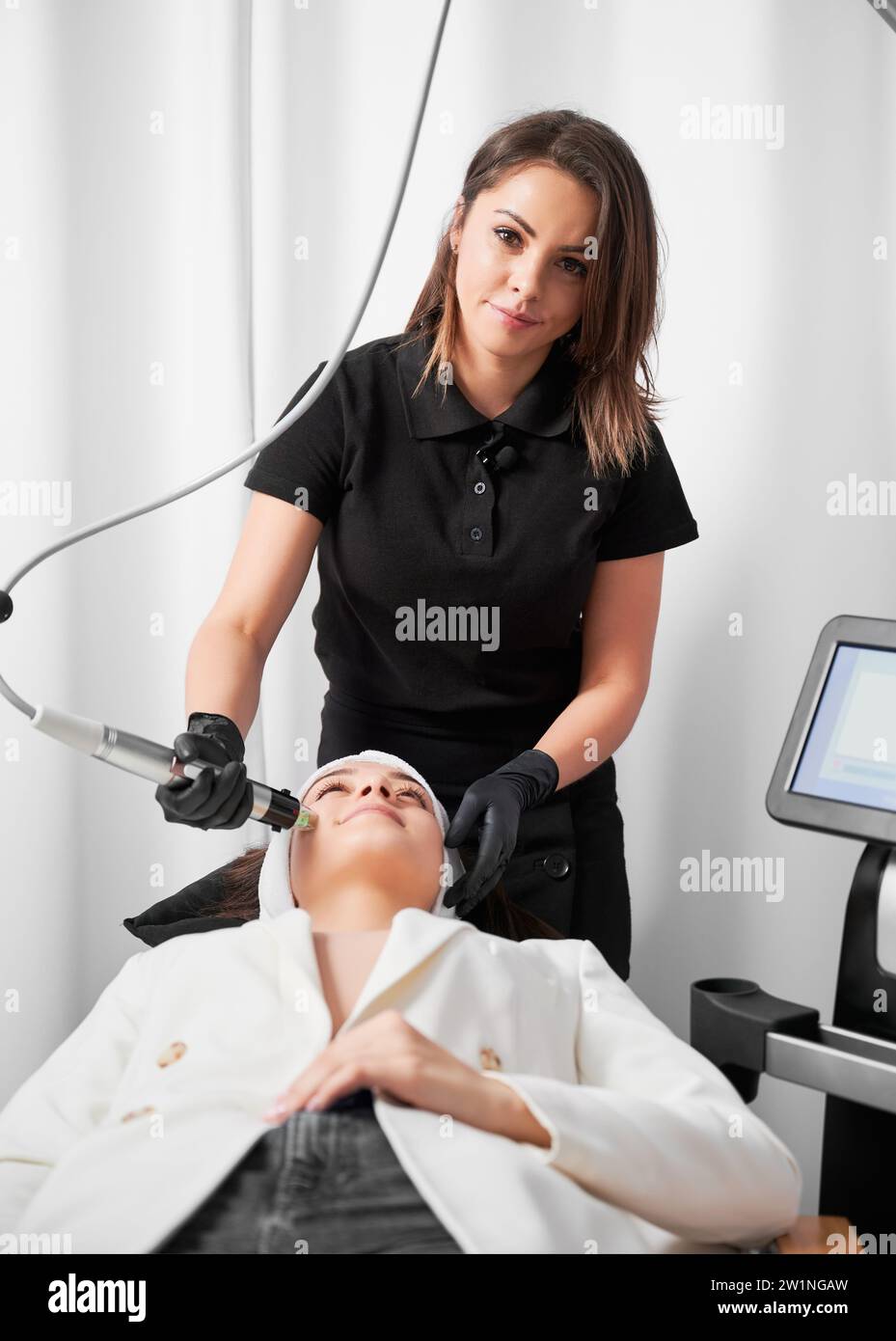 Medico estetista che utilizza attrezzature cosmetologiche professionali durante l'esecuzione della procedura di cura della pelle in un salone di bellezza. Cosmetologa femminile che cura il viso della donna con il dispositivo microneedling a radiofrequenza. Foto Stock