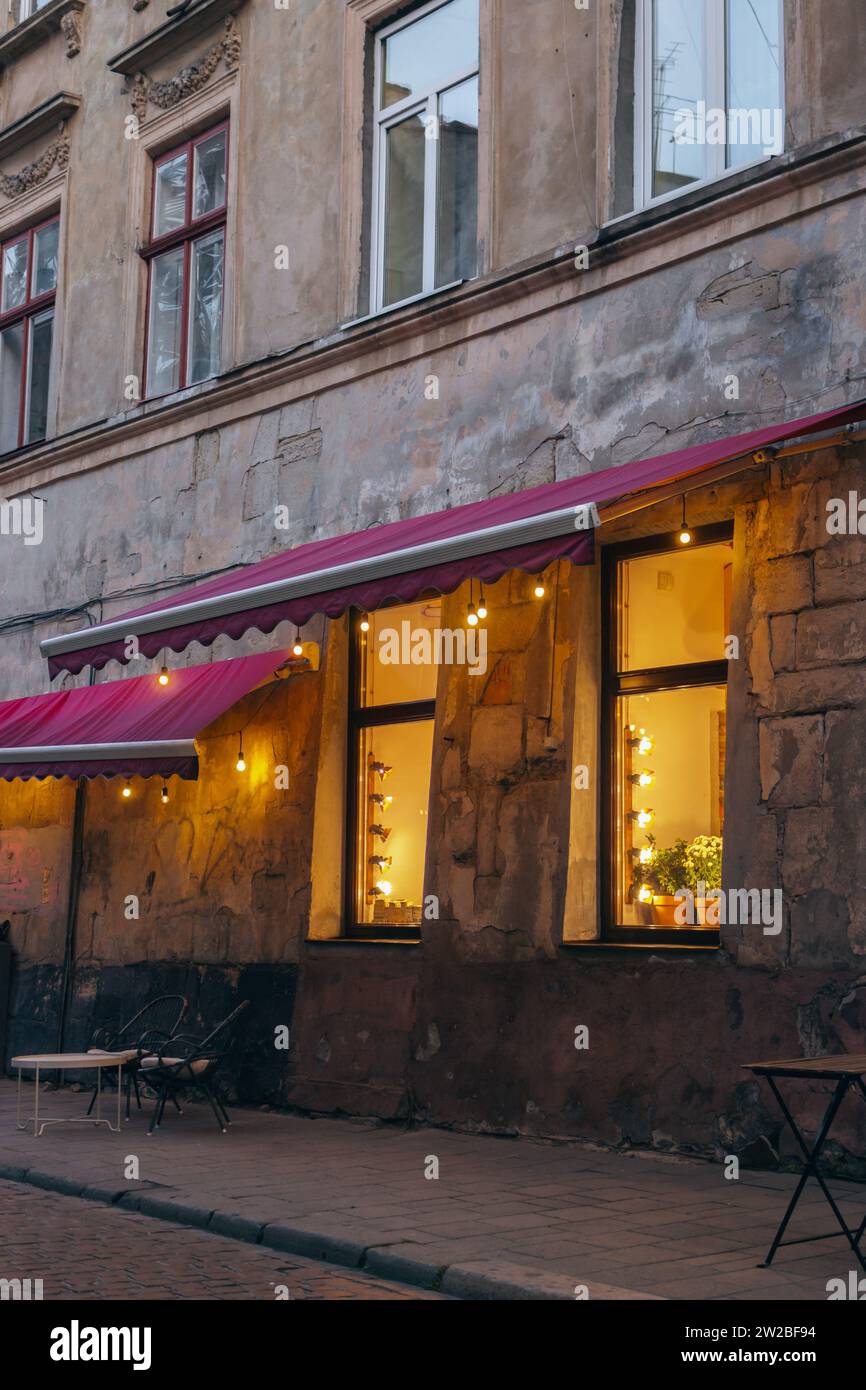 Luci calde nelle finestre del caffè nella vecchia città di Leopoli, Ucraina. Caffetteria con mobili da esterno e ghirlande nelle finestre. Caffè sul marciapiede in una casa antica Foto Stock