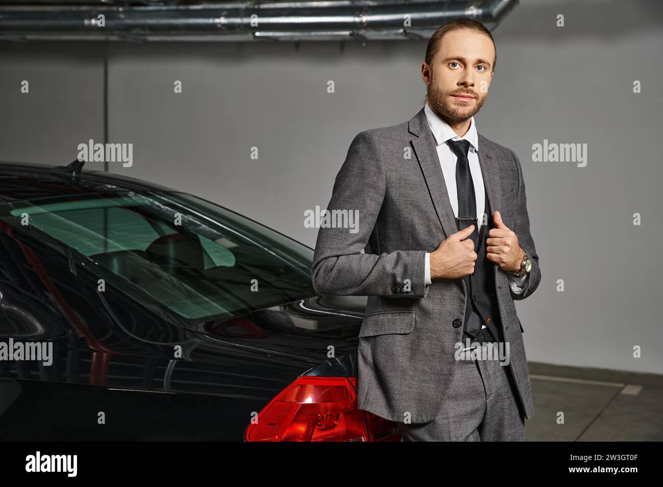 uomo attraente in tuta intelligente che guarda la fotocamera e posa davanti alla sua auto, concetto di business Foto Stock