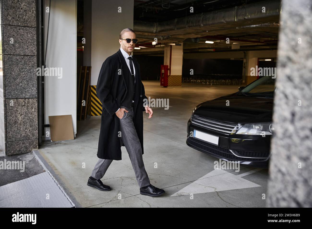 elegante uomo d'affari dall'aspetto gradevole con un cappotto nero con occhiali da sole che camminano e guardano la macchina fotografica Foto Stock