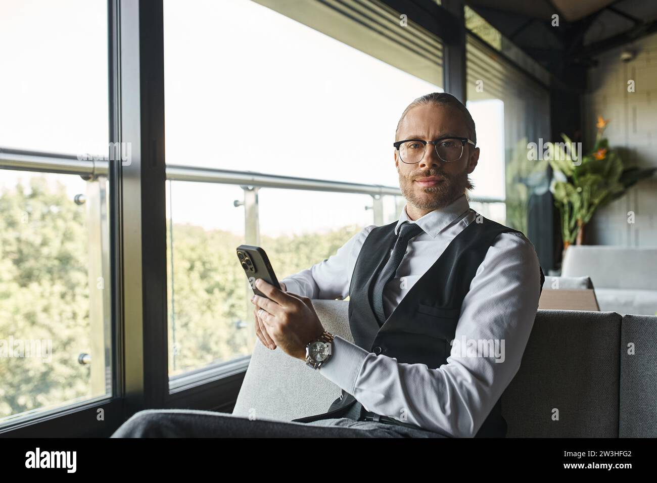 elegante uomo d'affari con un outfit elegante con occhiali che reggono il telefono e guardano la fotocamera Foto Stock