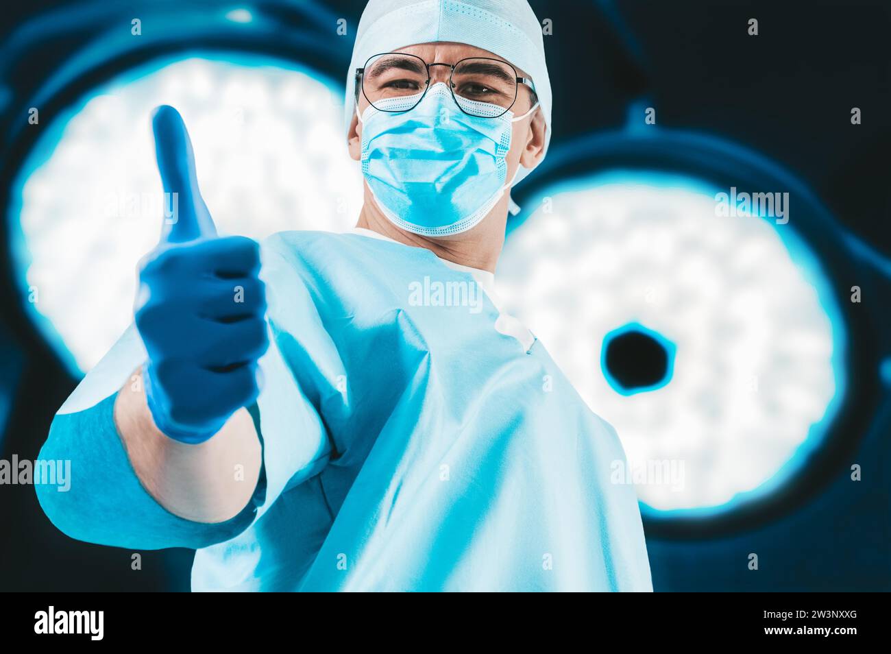 Il medico sullo sfondo delle lampade chirurgiche mostra un dito sollevato. Concetto di medicina. Supporti misti Foto Stock