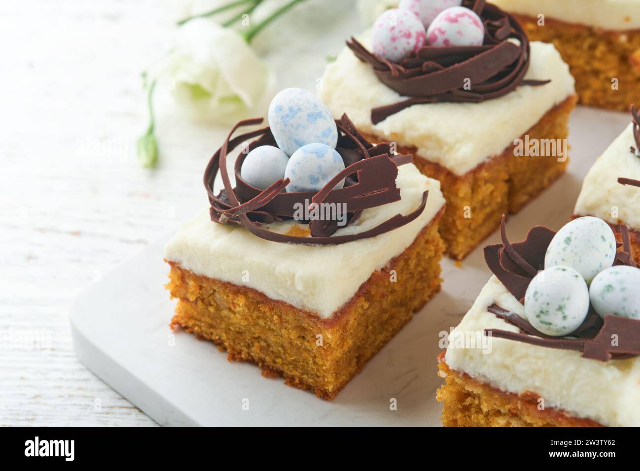 Barrette di carote pasquali decorate con nido di cioccolato e uova di caramelle al cioccolato fiorenti fiori di ciliegia o mela su sfondi rustici di legno chiaro. Foto Stock