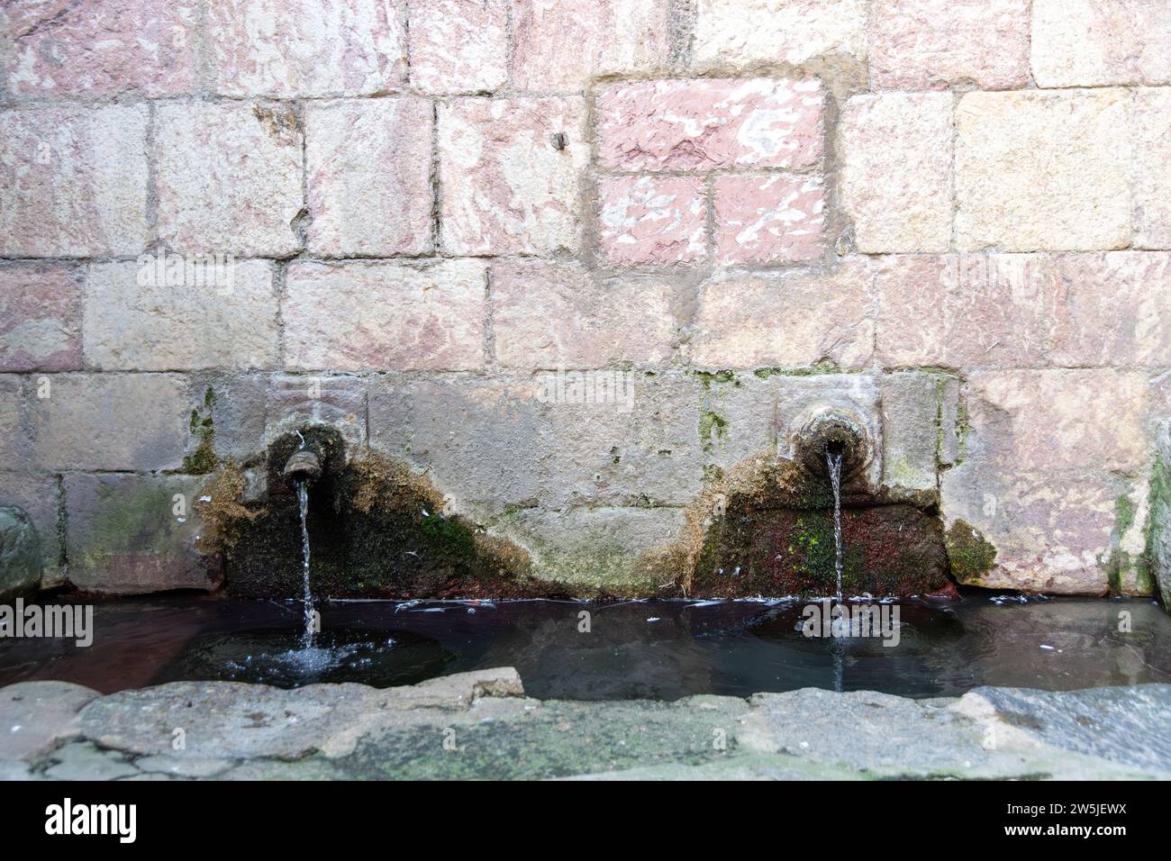 Due vecchie uscite d'acqua di una sorgente in un muro, da cui l'acqua scorre costantemente. Intorno ai rubinetti è possibile vedere le superfici ondulate sulla parete e a. Foto Stock