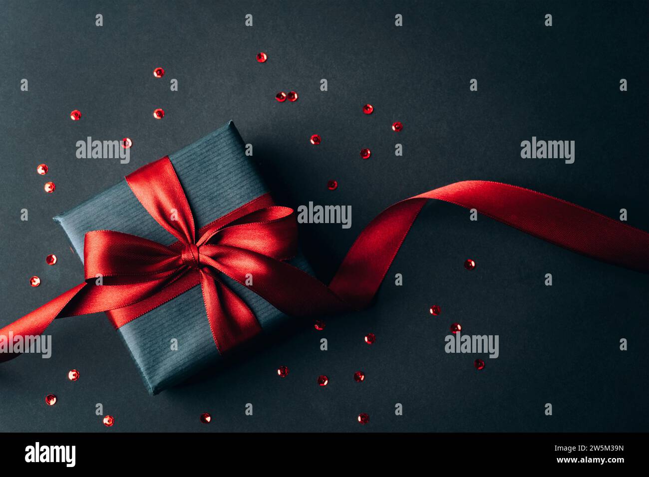 Confezione regalo con fiocco rosso su sfondo nero con coriandoli rossi. Natale, compleanno, festività. Vista dall'alto, base piatta. Foto Stock