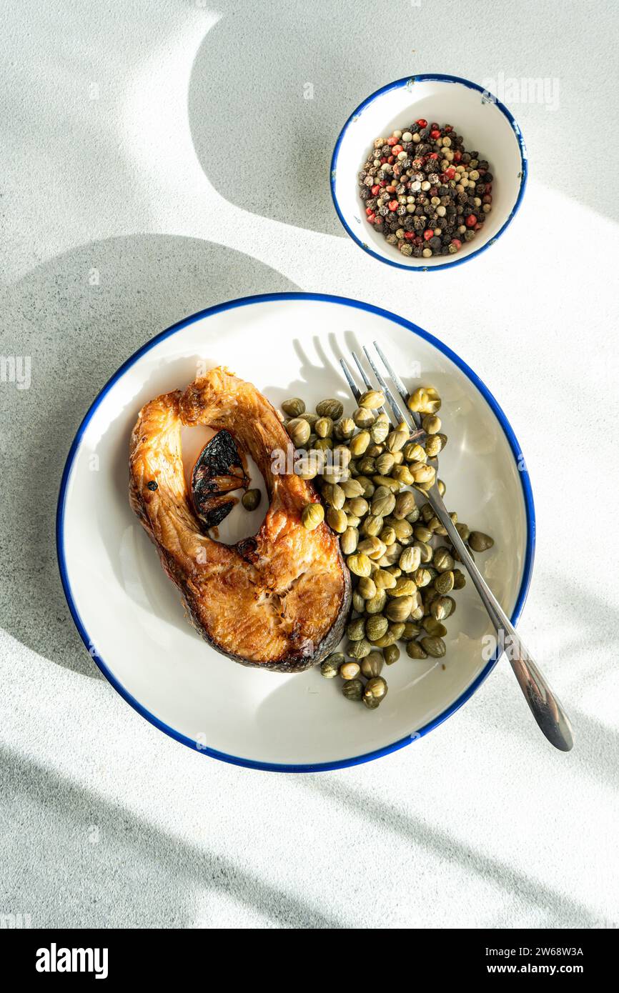 Vista dall'alto della bistecca di trota alla griglia ben preparata con capperi servita su un piatto bianco con forchetta posizionata su una superficie grigia vicino al recipiente con capperi Foto Stock