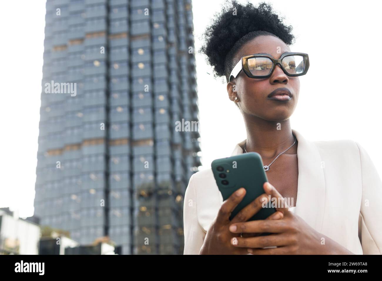 Giovane dirigente donna di colore con lo smartphone che guarda lontano mentre si trova contro l'edificio in città Foto Stock