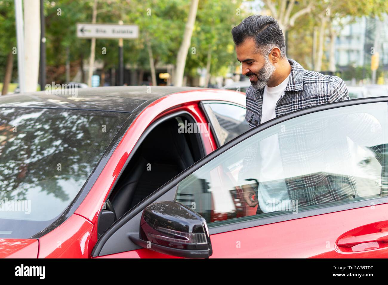 Imprenditore indiano positivo che entra in un'auto rossa con una porta aperta sul sedile del conducente sulla strada cittadina Foto Stock