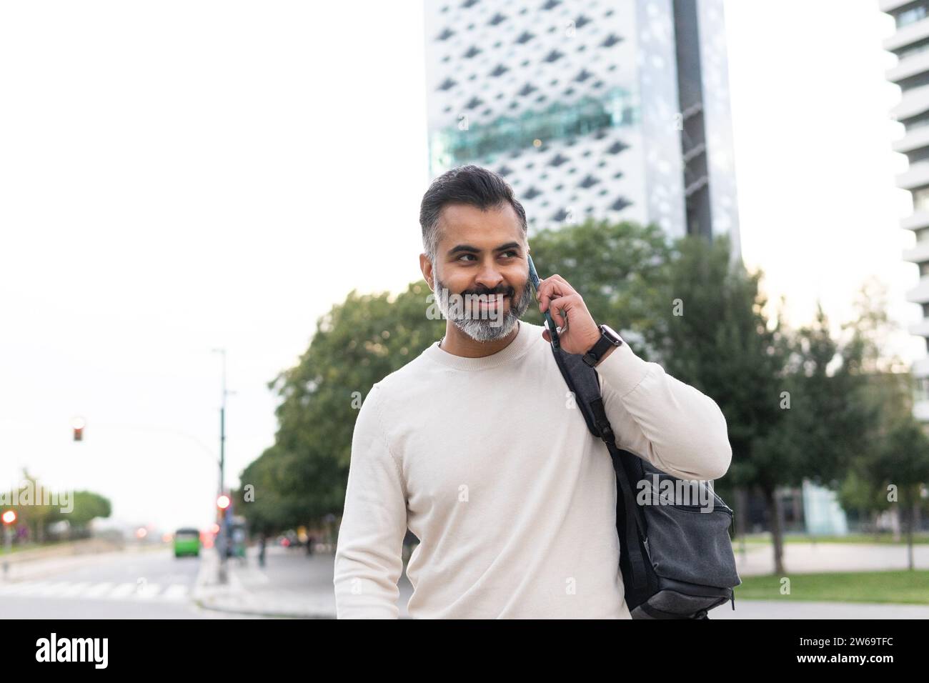 Uomo d'affari indiano barbuto positivo con zaino sorridente e che parla sullo smartphone su sfondo urbano sfocato Foto Stock