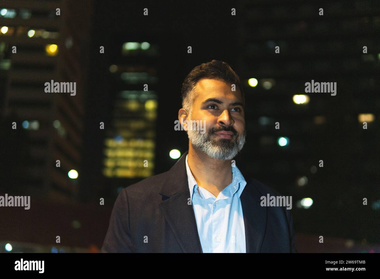 Un uomo d'affari etnico concentrato che indossa abiti eleganti contro lampade luminose e guarda fuori la sera Foto Stock